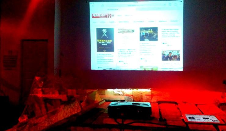 A dark room with a projection of a site or twitter feed titled protestify on a screen