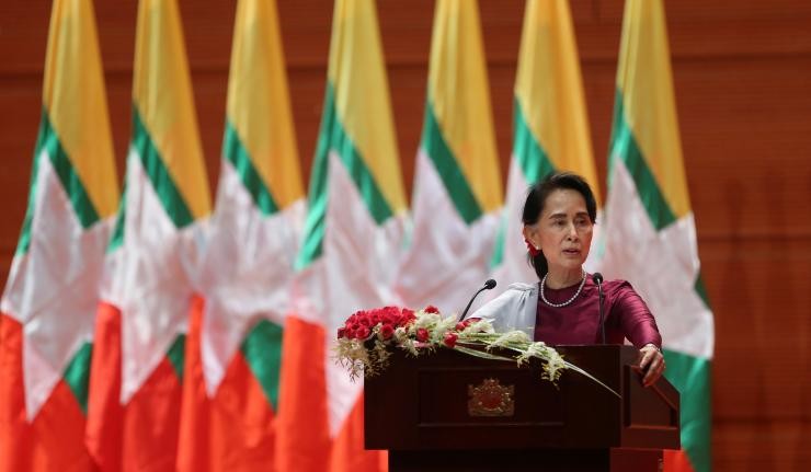 Aung Sang Suu Kyi speaking from a podium with Myanmar flags behind her