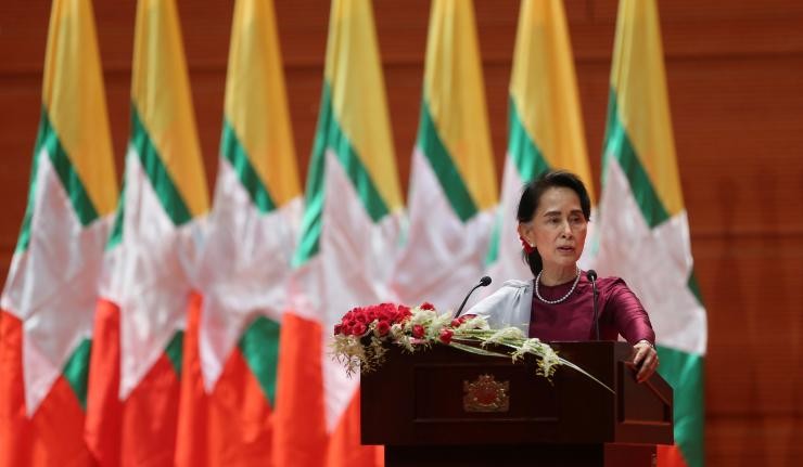 Aung Sang Suu Kyi speaking from a podium with Myanmar flags behind her Aung Sang Suu Kyi speaking from a podium with Myanmar flags behind her